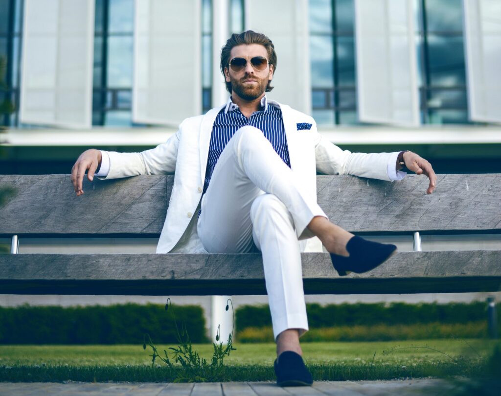 Fashionable man in white suit sitting confidently on a bench outdoors, embodying elegance and modern lifestyle.